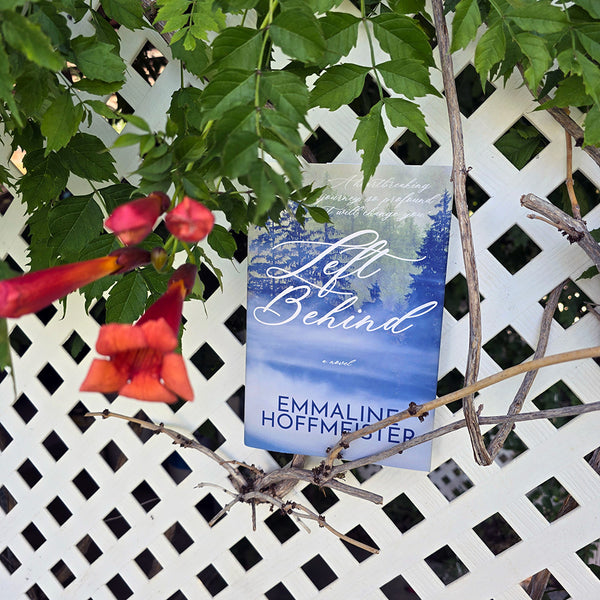 Book titled 'Left Behind' by Emmaline Hoffmeister with a scenic cover, placed among Honeysuckle greenery and flowers.